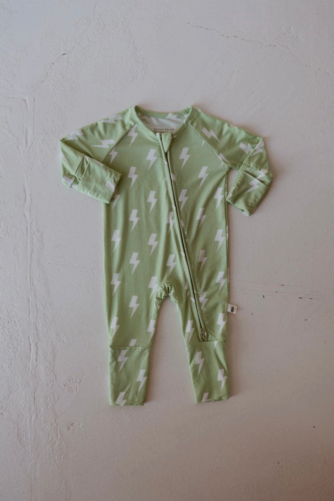 The forever french baby Bamboo Zip Pajamas | Flash is a light green, long-sleeve baby onesie with white lightning bolt patterns and a front zipper, crafted from breathable fabric and shown laid flat on a light textured surface.