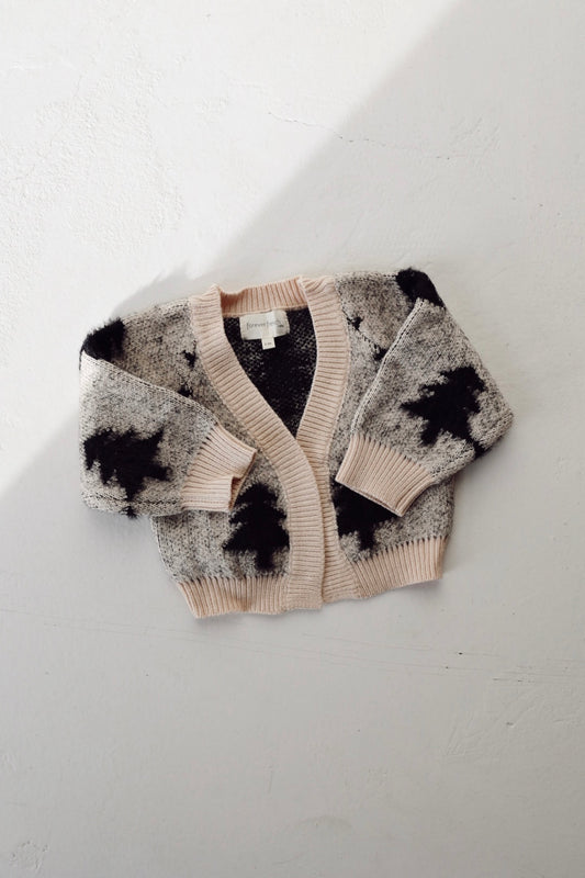 A soft, knitted "Silent Night" cardigan from forever french baby showcases a light pink trim and black abstract patterns, embodying charming elegance. It is displayed on a textured surface, illuminated by gentle lighting that accentuates its cozy vibe—an ideal addition to the Cozy Cardigan collection.