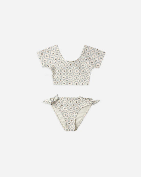 Introducing the Cropped Rashguard Set | Green Fleur from Rylee + Cru—a two-piece women’s swim set featuring a short-sleeve crop top and high-waisted bottoms, both adorned with a light green geometric Fleur pattern on a white background. This stylish rashguard swimwear provides UPF 50+ sun protection, so you can enjoy the water with added confidence.