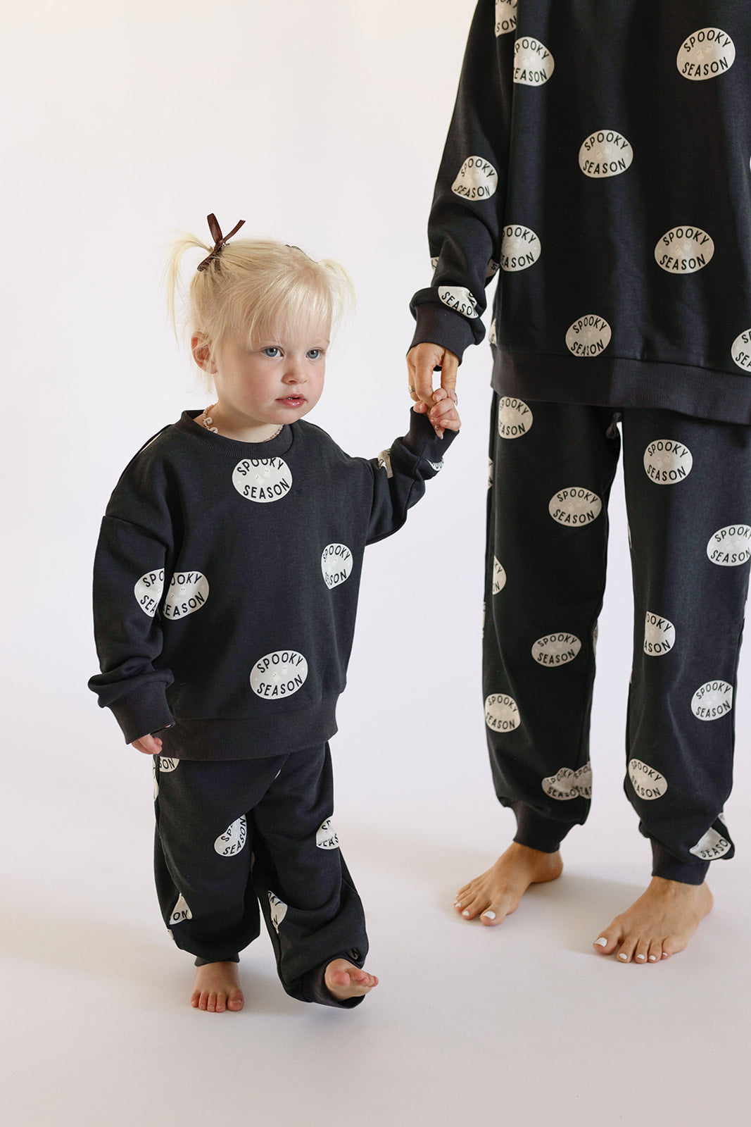 A toddler and adult, both barefoot, wear matching black "Child Sweat Set | Spooky Season" sets by forever french baby, featuring white circular patterns. The toddler holds the adult's hand as they stand together against a plain white background.