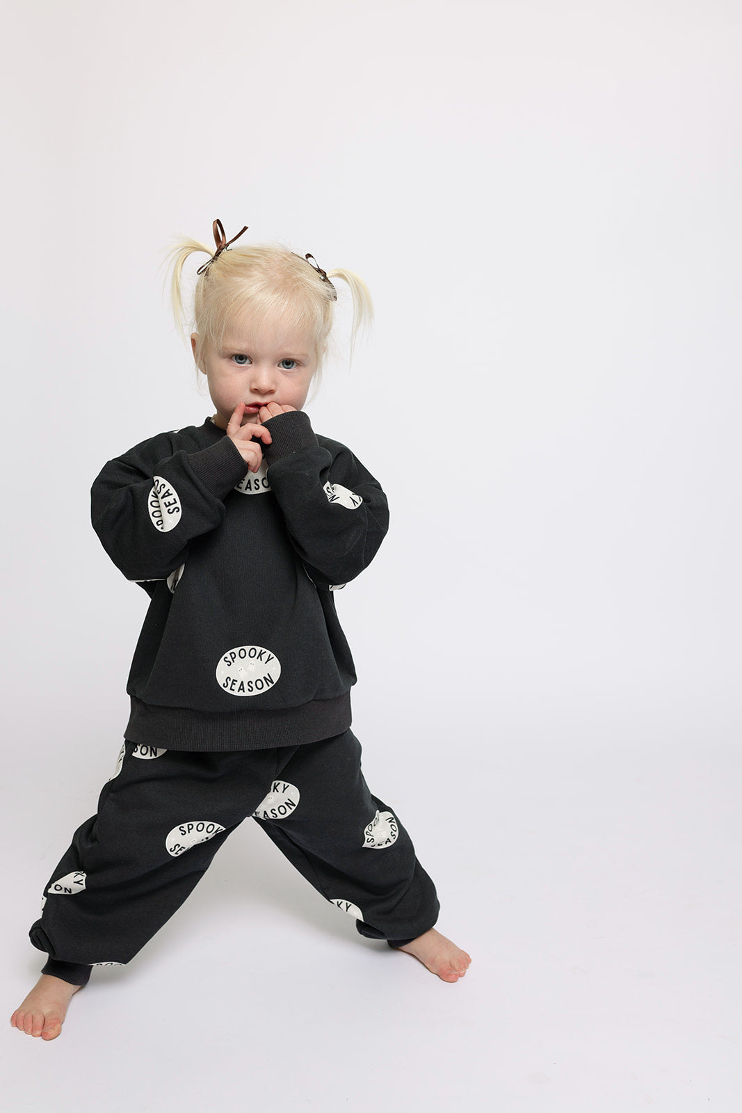 A young child with blonde pigtails stands against a white background in the forever french baby Child Sweat Set | Spooky Season—matching black sweatshirt and pants, both featuring "SPOOKY SEASON" in bold white ovals.