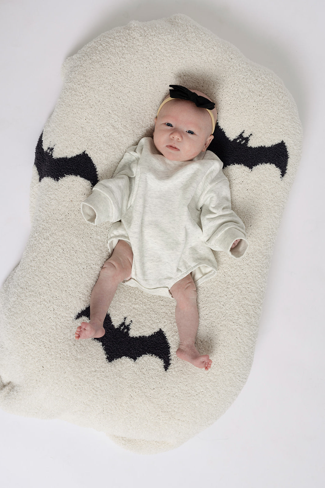 A baby in a cozy Heather Grey Sweatshirt Romper by forever french baby lies on a plush white mat with black bat shapes, looking up with relaxed arms and legs—perfect for family matching outfits or adorable newborn photos.