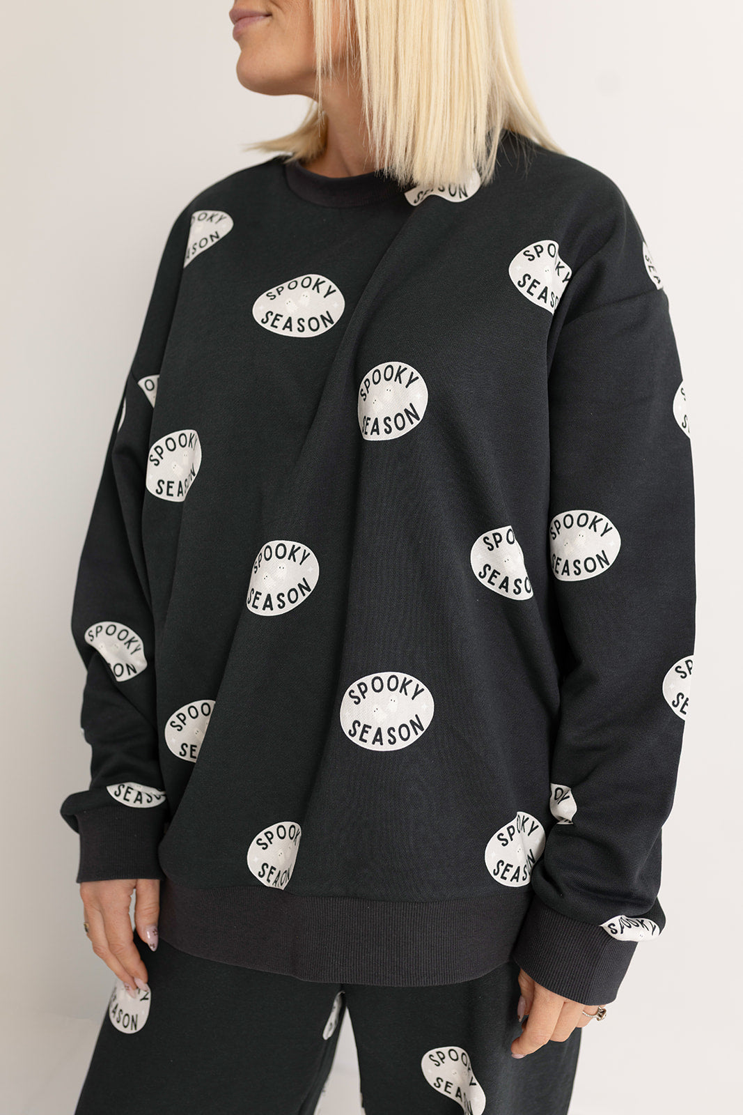 A woman with straight blonde hair wears the forever french baby Women's Sweat Set | Spooky Season—featuring a black sweatshirt and matching pants with repeating "Spooky Season" text. Her face is partially out of view.