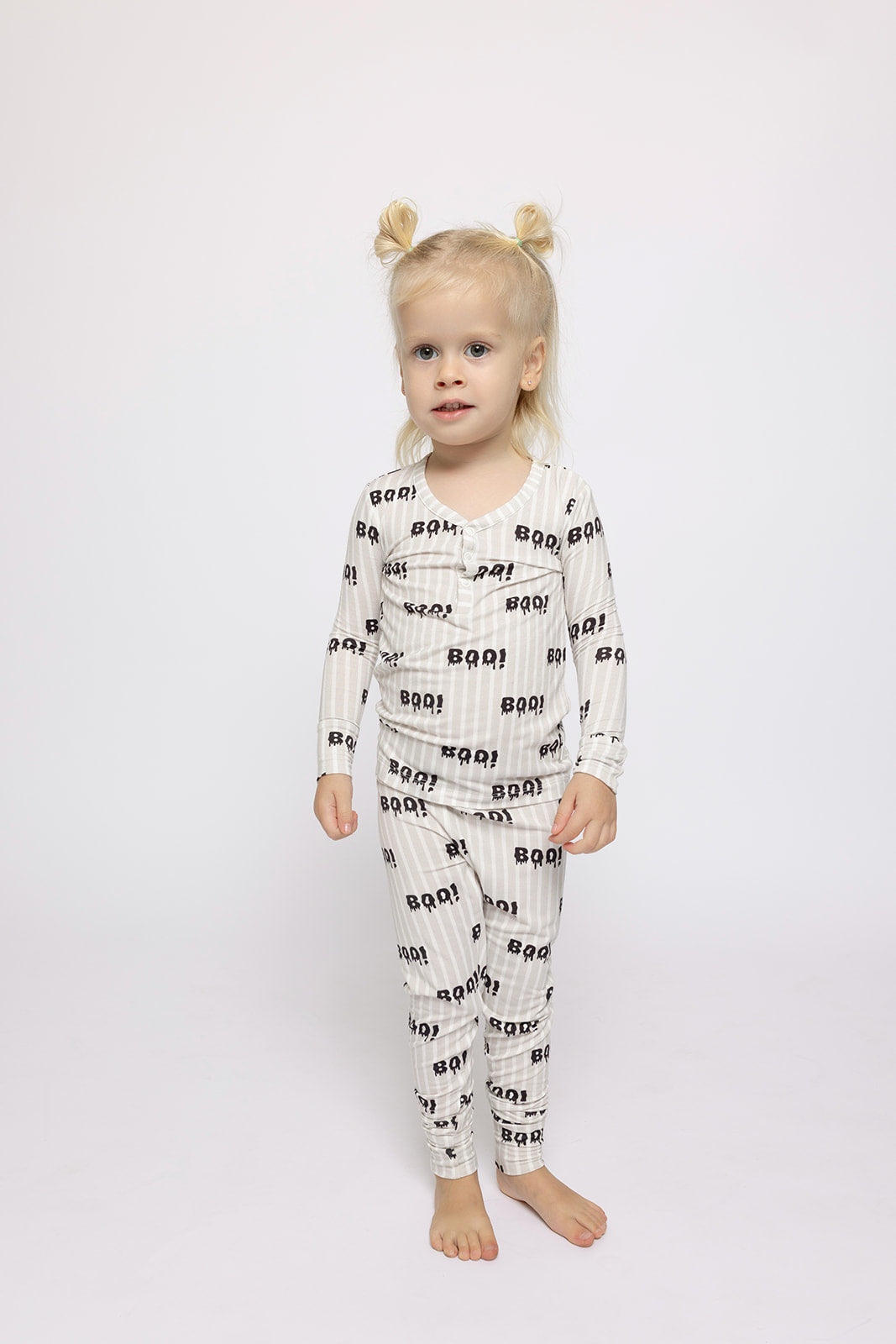 A young child with blonde hair in two buns stands barefoot on a white background, wearing forever french baby's Bamboo Two Piece Pajamas | Boo!—breathable children’s sleepwear featuring black "BOO!" prints for comfort and style.