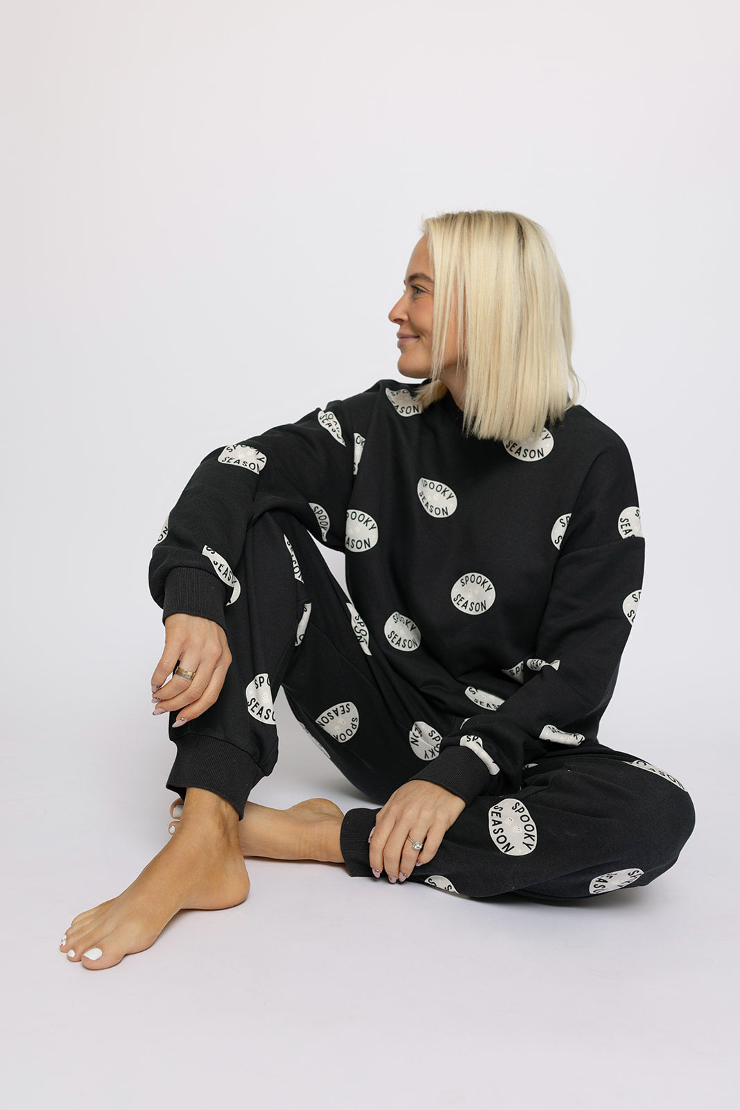 A barefoot blonde woman sits cross-legged, smiling to her left. She wears the forever french baby Women's Sweat Set | Spooky Season—black with white circular patterns—making a perfect spooky season outfit against a plain white background.
