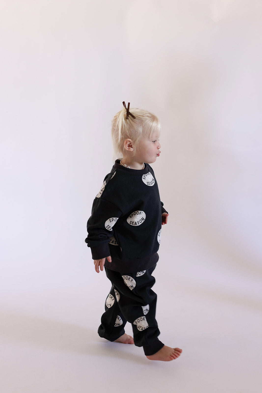 A blonde child with a hairpin walks barefoot on a light background, wearing the forever french baby "Spooky Season" Child Sweat Set—dark fabric with white circular patterns and text.