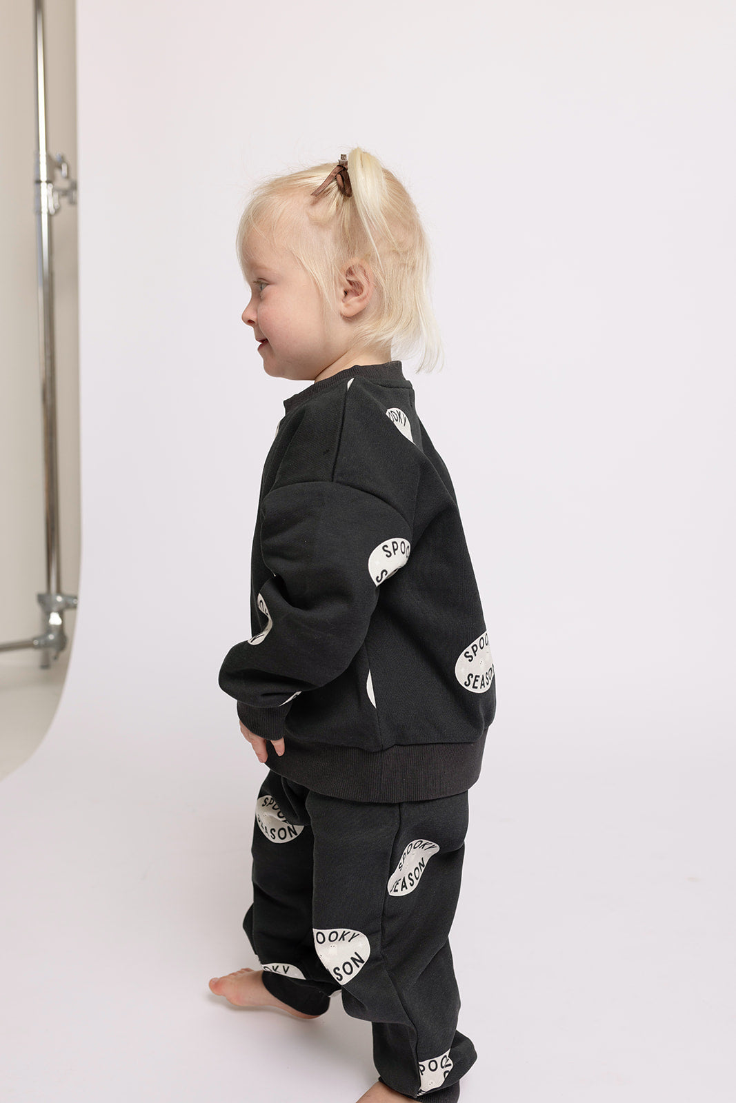 A young child with blonde hair models the forever french baby Child Sweat Set | Spooky Season—matching black sweatshirt and pants with white circular patches—standing barefoot on a white background, facing sideways.