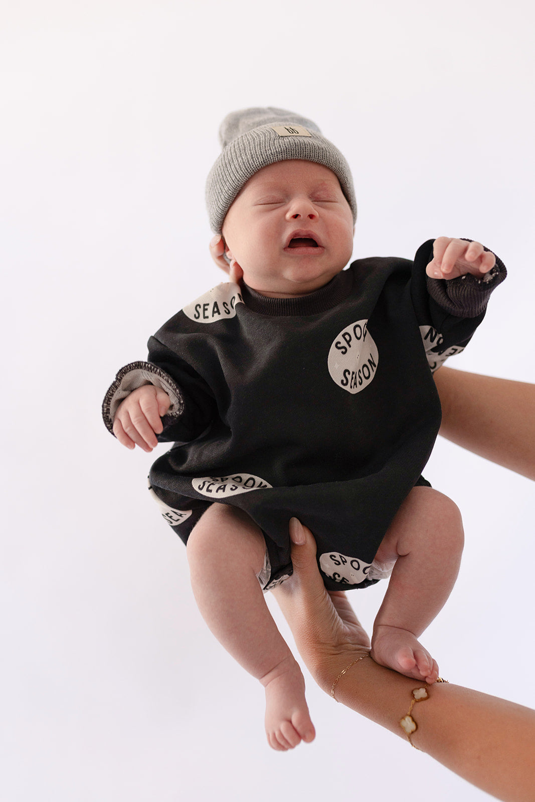 A baby in a gray beanie and forever french baby’s "Spooky Season" Sweatshirt Romper is held up by an adult, eyes closed and mouth open as if crying or squinting.