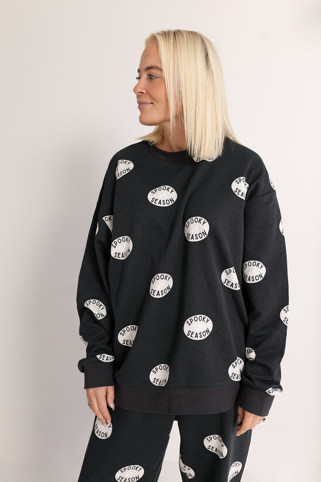 A woman with straight blonde hair wears the forever french baby Women's Sweat Set | Spooky Season—black with white "Spooky Season" ovals. She stands against a light background, smiling and looking left.