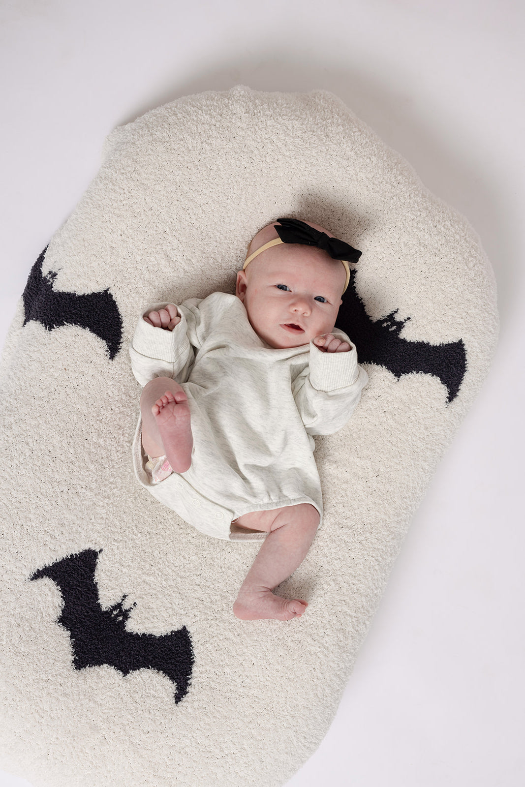 A baby in the forever french baby Sweatshirt Romper | Heather Grey lies on a soft, off-white cushion with black bat shapes, wearing a black bow headband. The background is white.