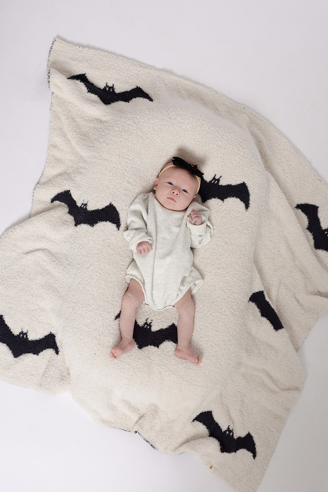 A baby wearing the forever french baby Sweatshirt Romper in Heather Grey lies on a soft cream blanket with large black bat silhouettes, set against a plain white background.