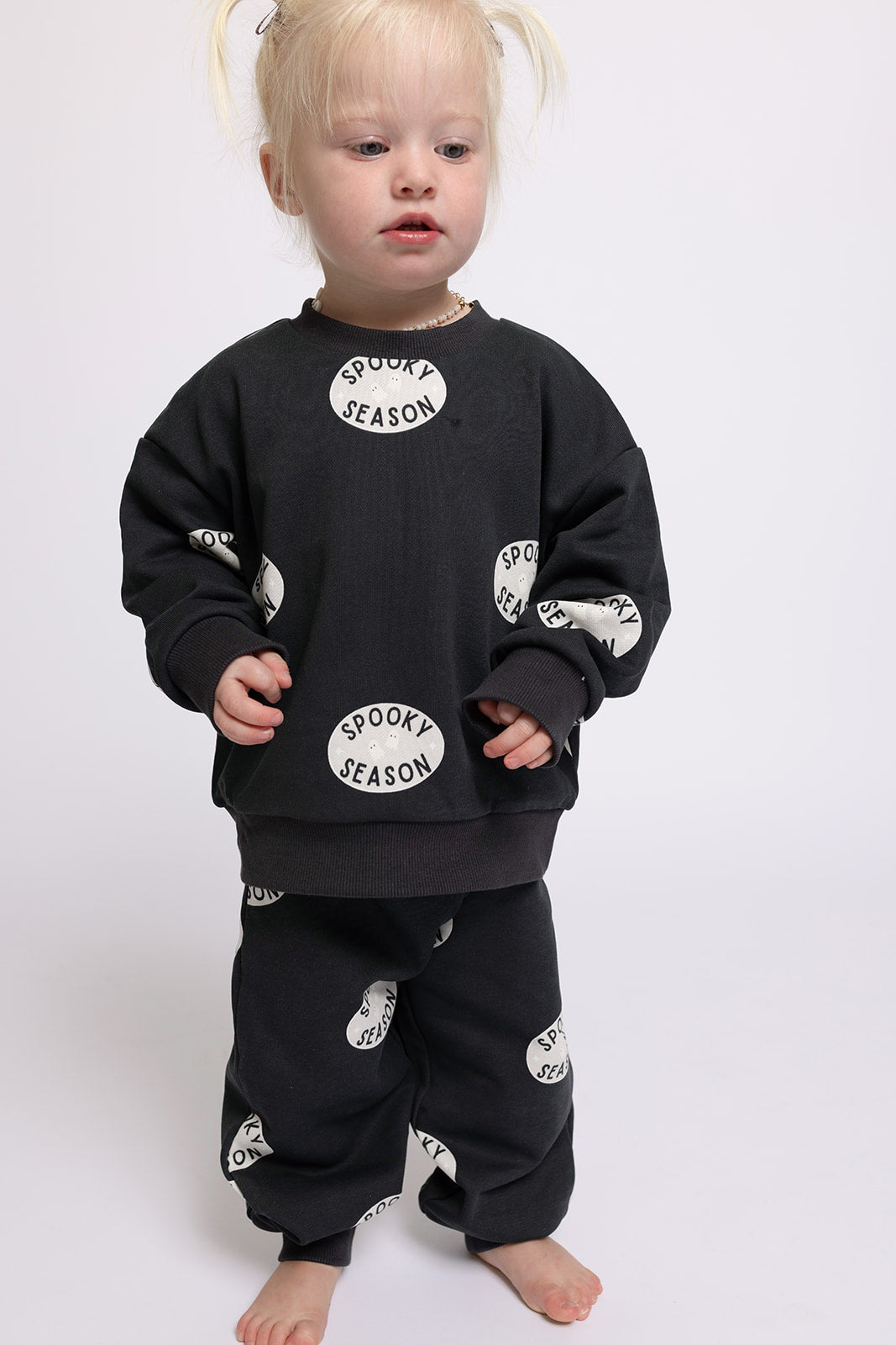 A blonde child in pigtails models the forever french baby "Child Sweat Set | Spooky Season," featuring white circle lettering, standing barefoot against a plain backdrop—ideal for matching with a mom set.