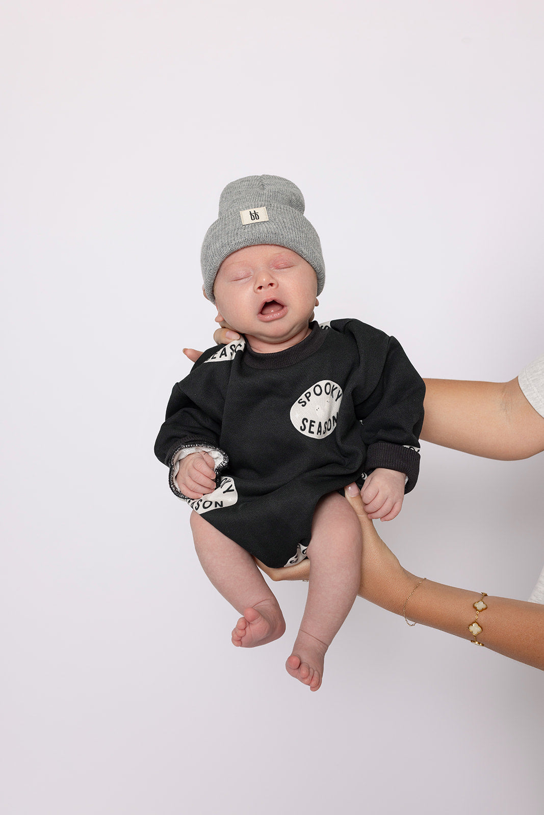 A baby, eyes closed and mouth slightly open, is gently held by an adult while wearing the forever french baby "Spooky Season" Sweatshirt Romper and a grey knit hat against a plain background.
