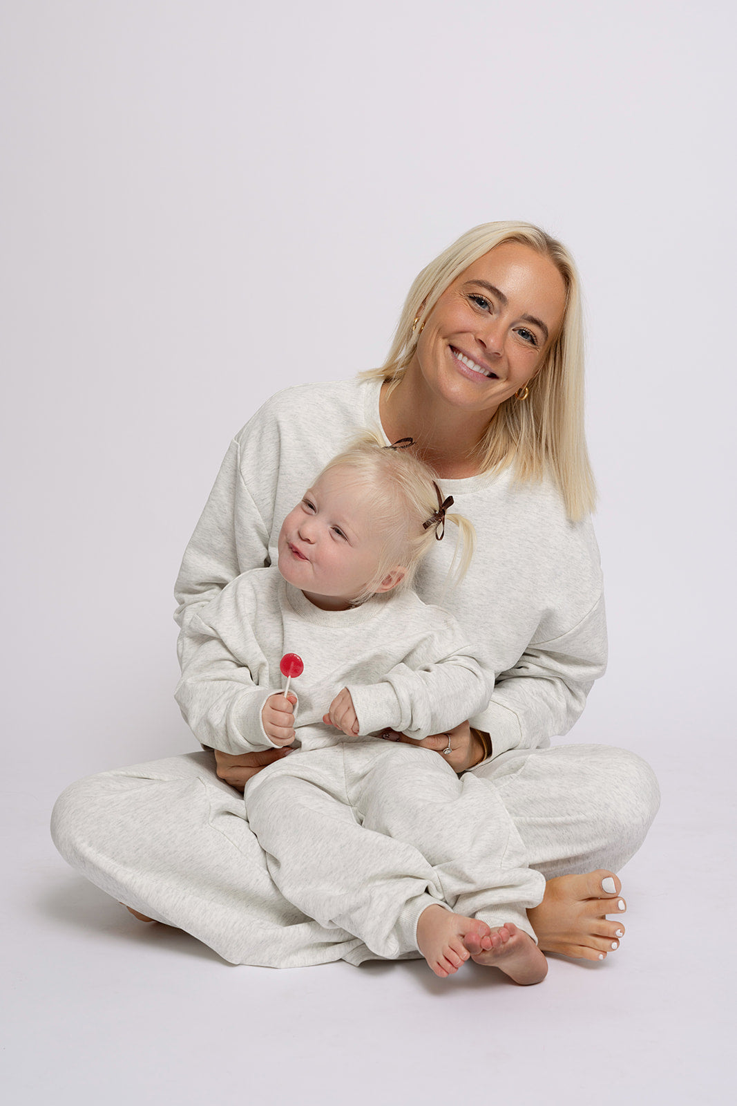 A smiling woman sits with a young girl in her lap, both wearing matching forever french baby Child Sweat Set in Heather Grey. The girl, with pigtails and a red lollipop, poses against a plain white background—perfect for cozy family looks.