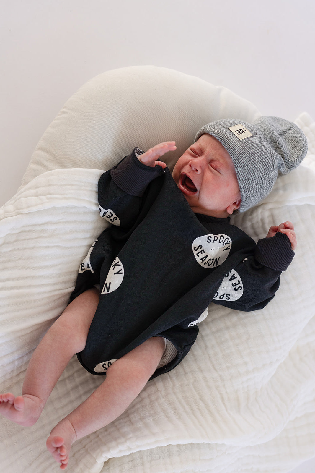 A baby in forever french baby's Sweatshirt Romper | Spooky Season, featuring white circular text patterns, and a gray beanie lies on a soft white blanket—an adorable, cozy Halloween outfit for your little one.