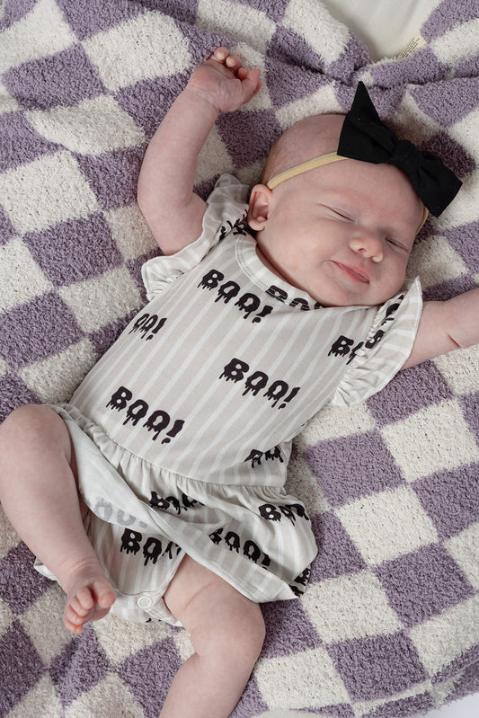 A baby in a forever french baby Bamboo Ruffle Romper | Boo! with a black bow headband sleeps peacefully on a purple and white checkered blanket, one arm raised above their head.