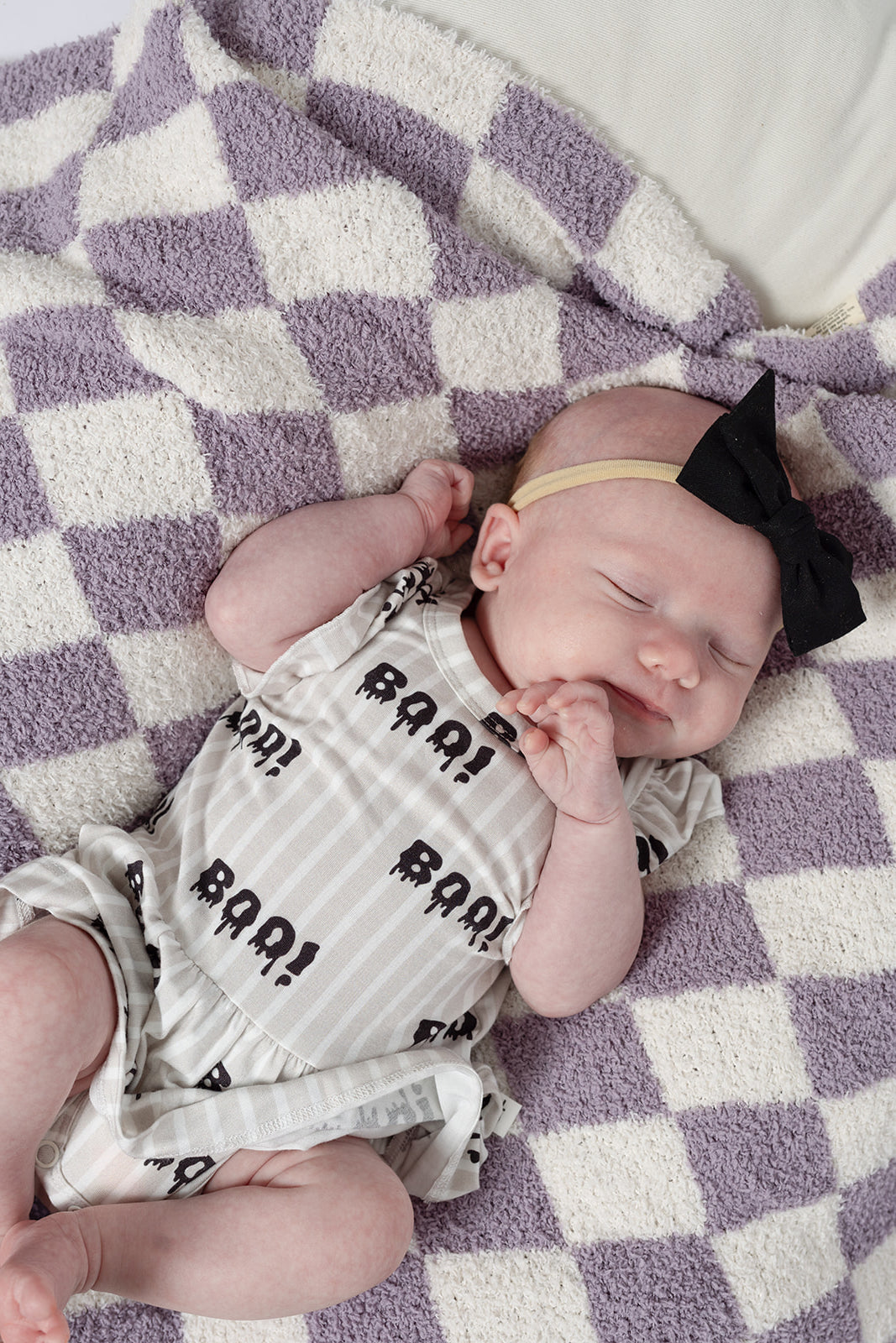 A sleeping baby in a "BAA!" onesie and black bow headband rests on the forever french baby Plush Blanket | Grape Soda, an Oeko-Tex certified, purple and white checkered soft blanket.