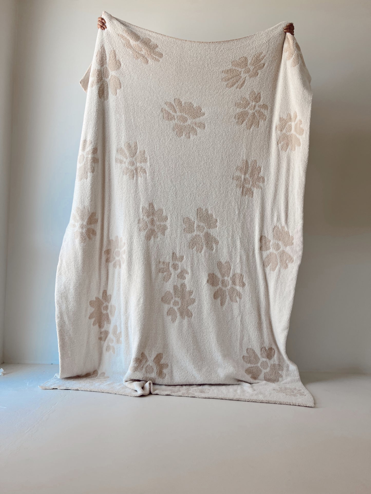 The Plush Blanket | Lazy Daisy from forever french baby, featuring a cream-colored design adorned with large, light brown flowers, is held up against a plain white wall. Crafted from cozy Microfiber feather yarn, the blanket drapes elegantly with soft folds at the bottom as two hands grasp its top corners to keep it in place.