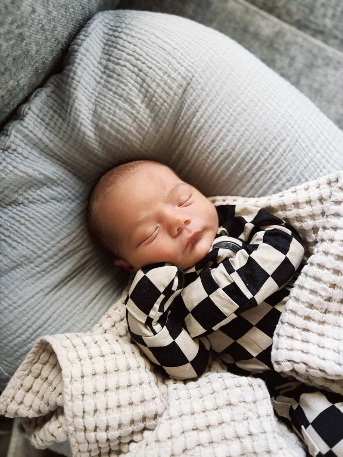 A newborn sleeps peacefully on a soft pillow, wrapped in a cream blanket and cozy forever french baby Bamboo Zip Pajamas in Black Checkerboard.