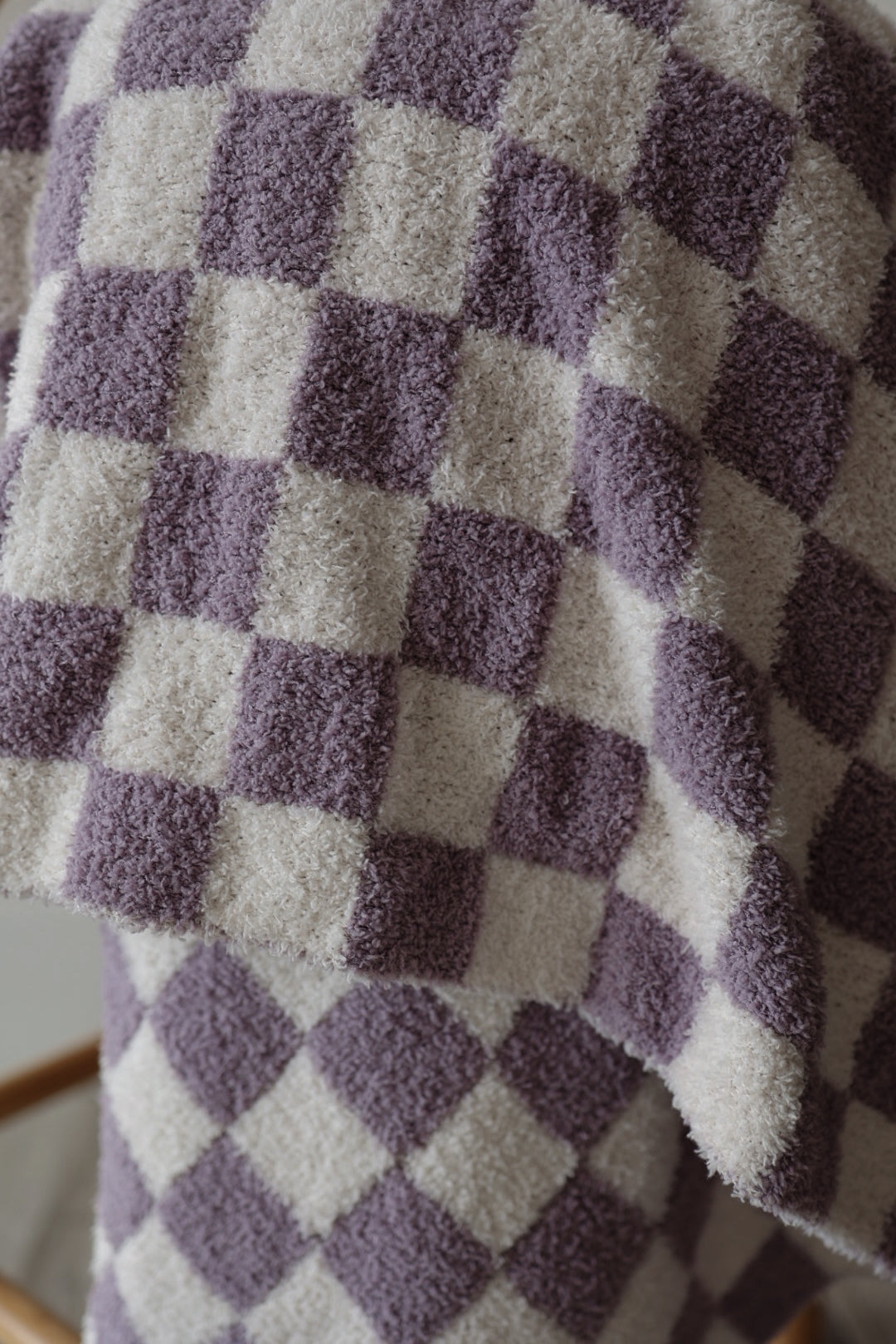 The forever french baby Plush Blanket | Grape Soda, with its soft, fuzzy texture and light purple checkered pattern, drapes elegantly over a wooden surface—bringing cozy comfort to any space.