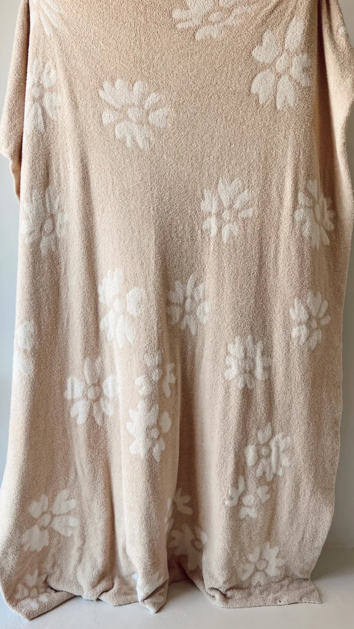 The Plush Blanket | Lazy Daisy from forever french baby is displayed hanging. This beige blanket, made from microfiber feather yarn and meeting OEKO Standard 100, boasts a soft, velvety-rich texture with a delightful pattern of white floral designs scattered across it. It looks exceptionally plush and cozy.