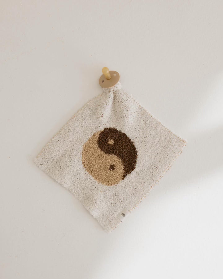 A white wall showcases a soft fabric square known as the Lovey | Brown Sugar Yin Yang from forever french baby, featuring a brown and beige yin-yang symbol in the center. The square is hanging by a beige pacifier holder attached to its top corner, blending functionality with charm.