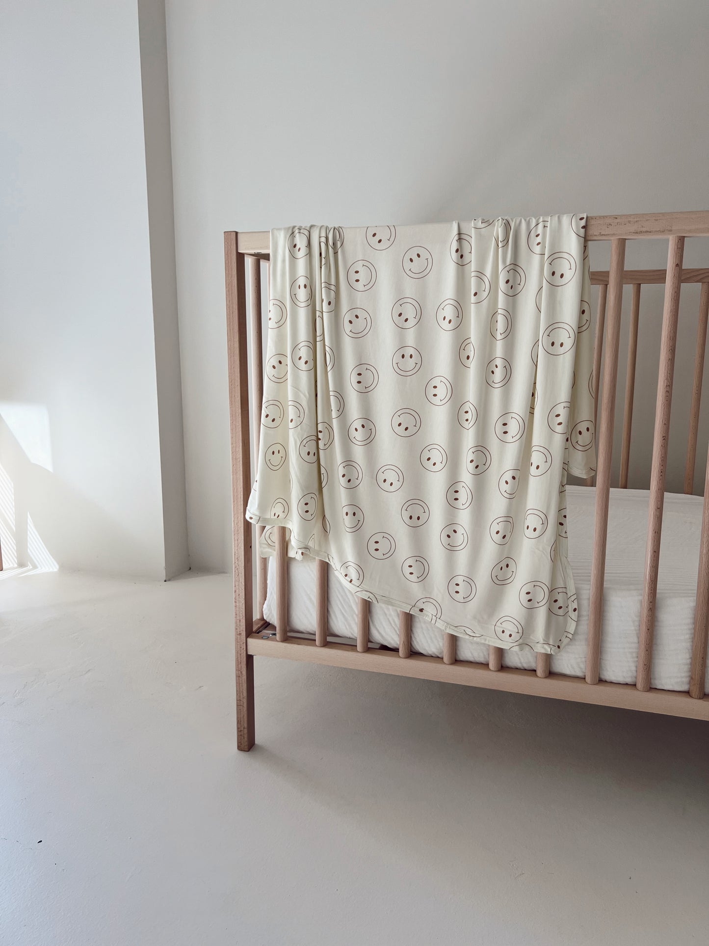 A minimalistic wooden baby crib is placed in a bright, light-filled corner of the room. An ivory "Just Smile" Bamboo Swaddle from forever French baby, featuring a cheerful smiley face pattern, is draped over the side of the crib. The room features soft, neutral tones, providing year-round comfort.