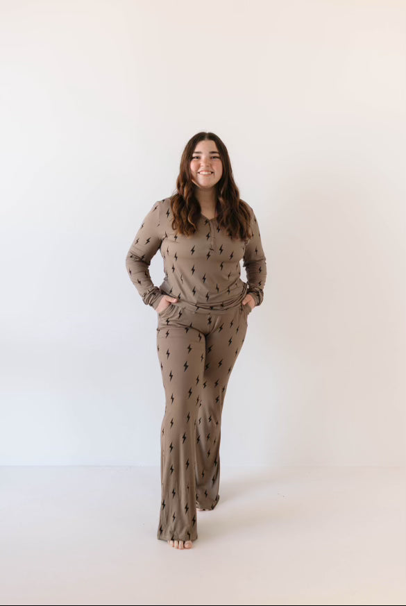 A person with long brown hair stands barefoot against a plain white background, wearing the Women's Bamboo Pajamas in Brown & Black Lightning Bolt by forever french baby. The beige lounge set, adorned with black lightning bolt patterns, is made from breathable fabric. They have their hands in their pockets and are smiling.