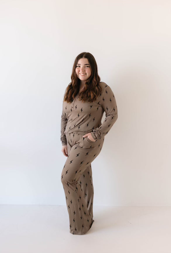A person with long, wavy hair stands against a plain white backdrop, smiling at the camera. They are wearing forever french baby's Women's Bamboo Pajamas in Brown & Black Lightning Bolt design, consisting of a long-sleeved top and matching pants, with one hand in their pocket.