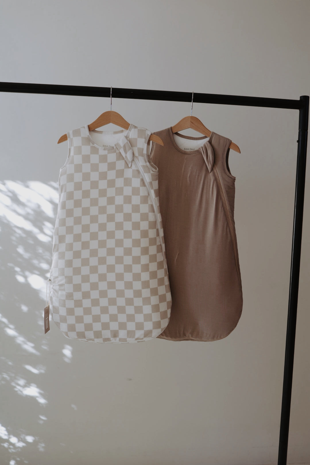 A Bamboo Sleep Sack by forever french baby hangs on a black clothing rack. One features a beige and white checkerboard design, while the grey version offers a solid look. Soft light and shadows highlight each wearable blanket against the plain wall.