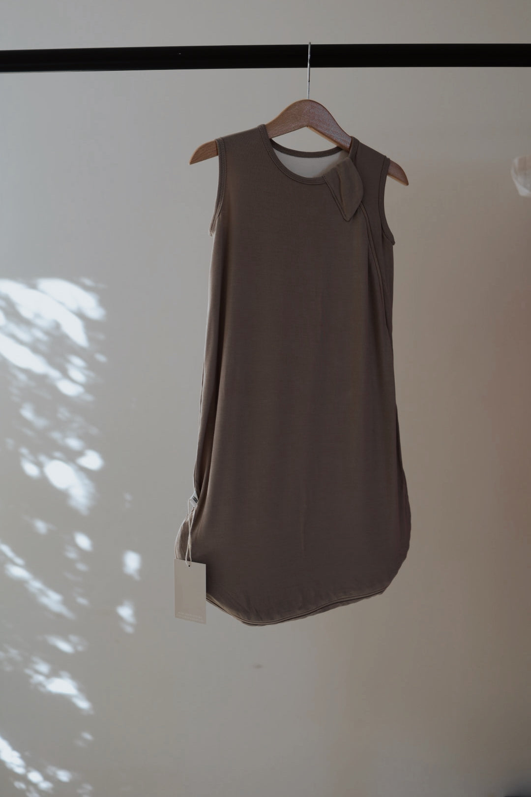 The forever french baby Bamboo Sleep Sack in Grey, featuring a sleeveless design and round neckline, hangs on a wooden hanger against a softly lit, plain wall with dappled shadows. A tag is attached.