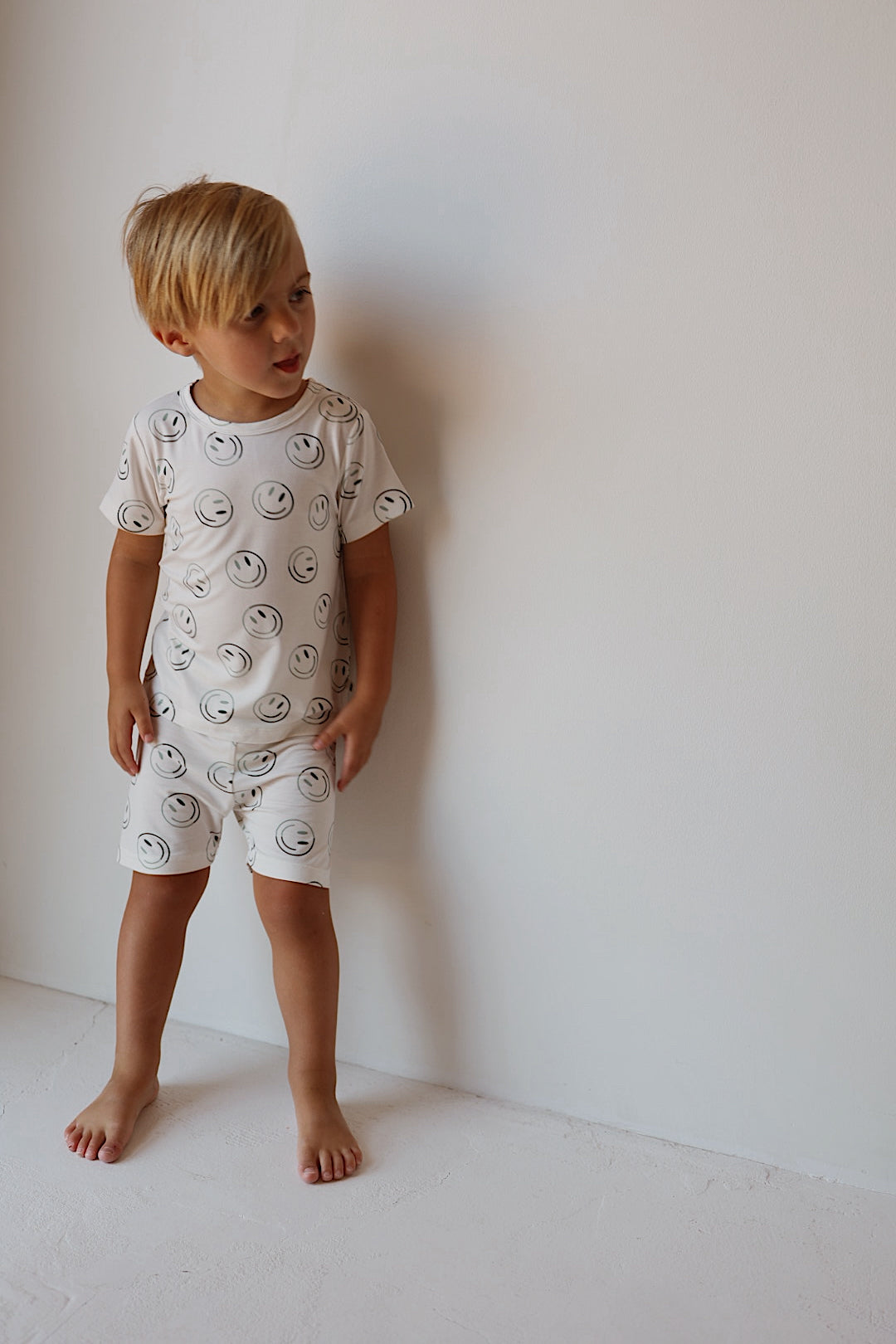 A young child with blond hair stands barefoot against a white wall, wearing the forever french baby Bamboo Short Set | Green Smiles—a hypoallergenic kids clothing option featuring smiley face patterns.