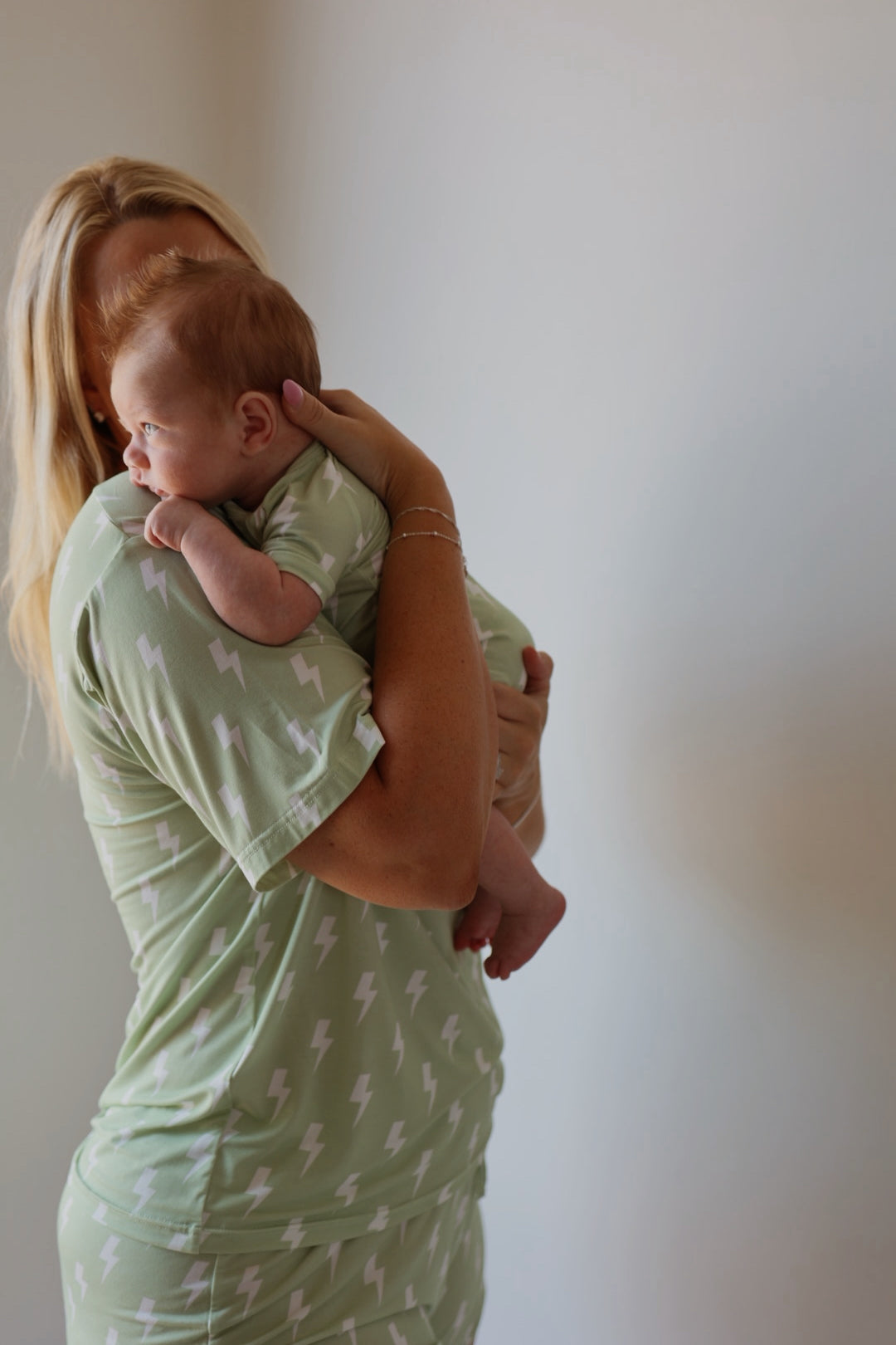 A woman with long blonde hair holds a baby against her shoulder. Both wear matching “Shortie Bamboo Zip | Flash” pajamas by forever french baby, featuring light green bamboo fabric and white lightning bolt patterns.