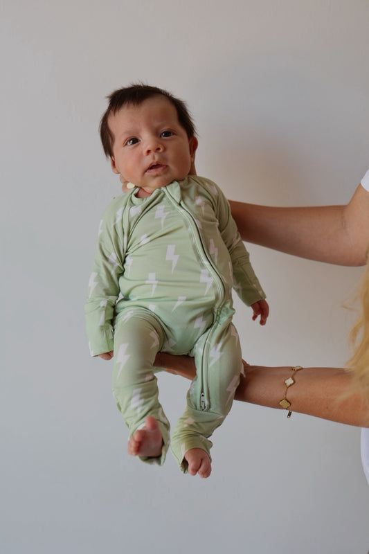 A baby in forever french baby's Bamboo Zip Pajamas | Flash—light green with white lightning bolts—is held upright by an adult against a plain light background.