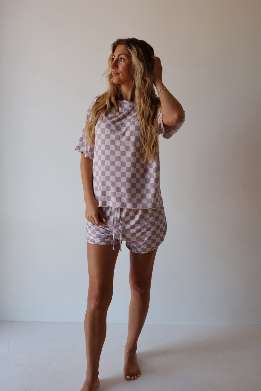 A woman with long wavy hair stands barefoot against a light background, wearing the forever french baby Women's Bamboo Short Set in Grape Soda—a matching lavender and white checkered loungewear set—while looking to the side and touching her hair.