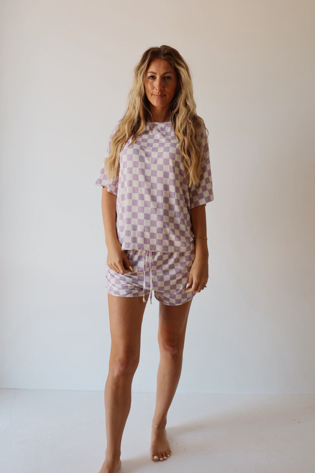 A woman with long, wavy hair stands barefoot against a plain background, wearing the forever french baby Women's Bamboo Short Set in Grape Soda—a lilac and white checkered ultra-soft bamboo loungewear set with a short-sleeve top and drawstring shorts.