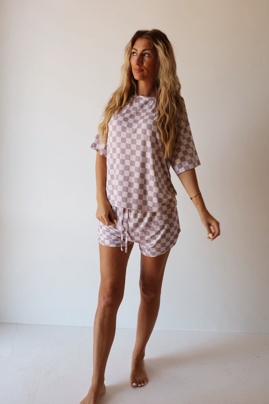 A woman with long wavy hair stands barefoot against a plain background, wearing the forever french baby Women's Bamboo Short Set in Grape Soda — a matching lavender and white checkered loungewear set.