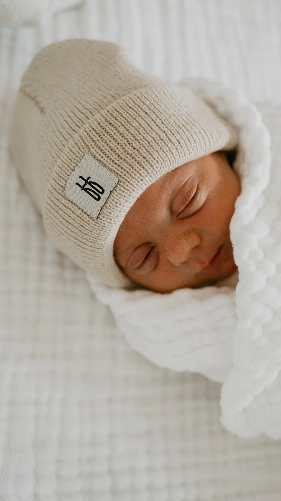 A newborn baby sleeps peacefully, wrapped in a white textured blanket and wearing an oatmeal-colored ff Knit Beanie from forever french baby. The baby is lying on a soft white surface, creating a serene and cozy scene.