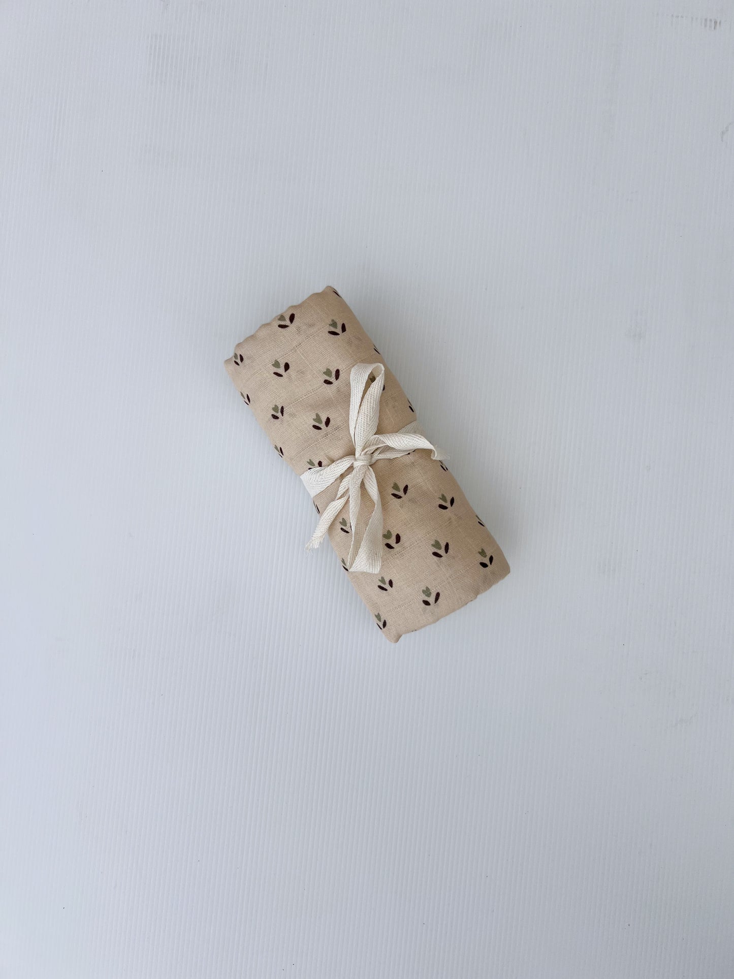 A cylindrical package wrapped in brown paper with black plant motifs and a white ribbon rests on a plain surface. Inside, find the ultra-soft Muslin Swaddle | Flora by forever french baby, crafted from 100% cotton for the ultimate comfort and care for your little one.