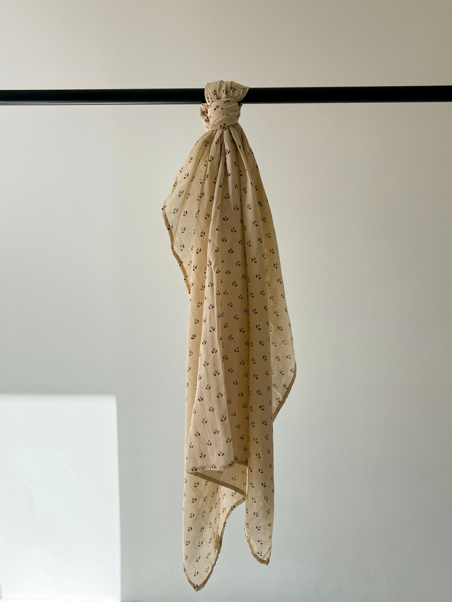 A Muslin Swaddle | Flora by forever french baby, with a 100% cotton texture, drapes gracefully from a black rod on a light-colored wall. Soft lighting casts a gentle shadow, highlighting this ultra-soft beige piece adorned with small black patterns.