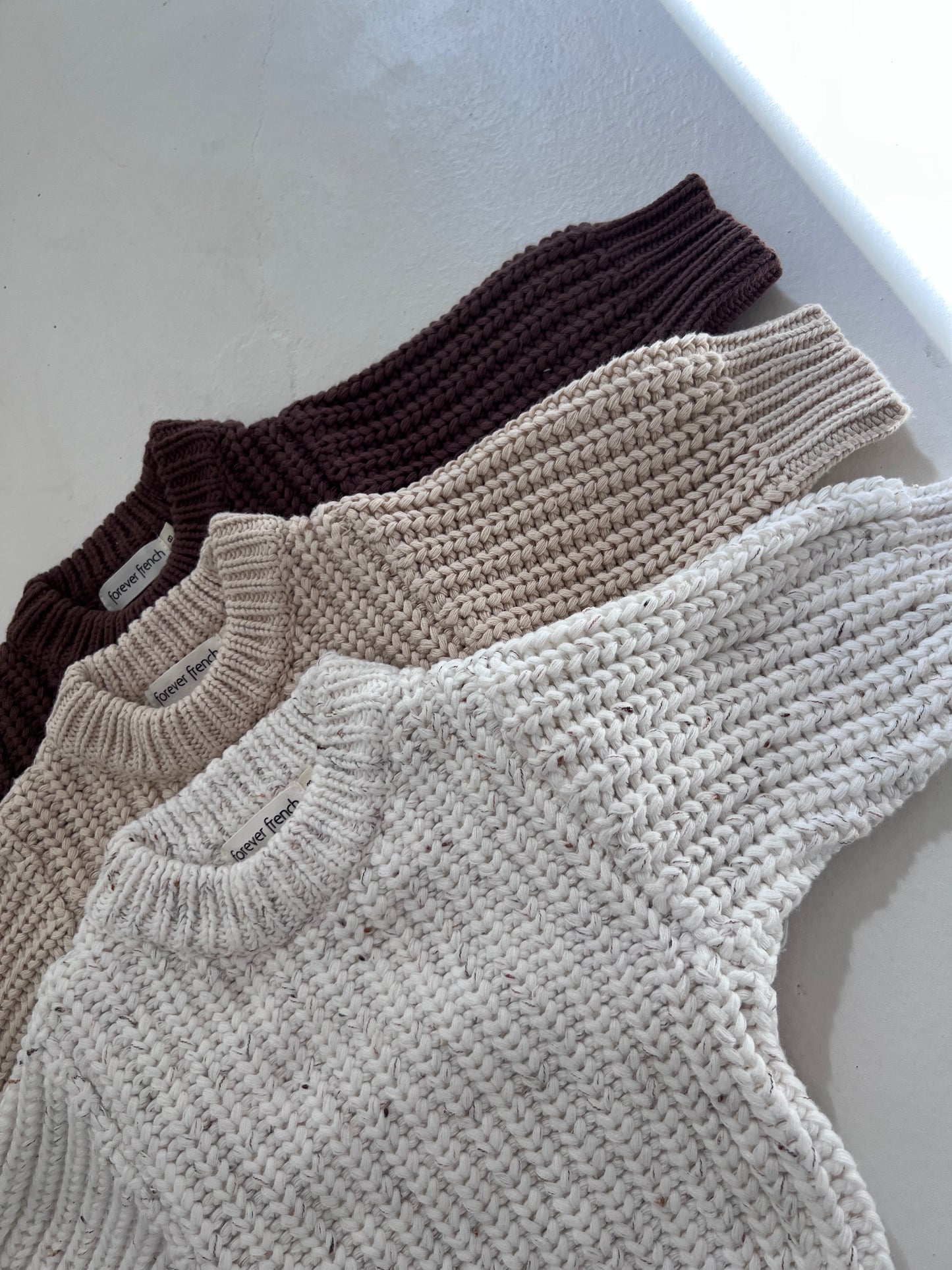 Four "Child Knit Sweater | Cloud Confetti" by forever french baby in cream, beige, and brown are stacked on a white surface. Each has a visible neckline label with detailed, prominent knit texture.