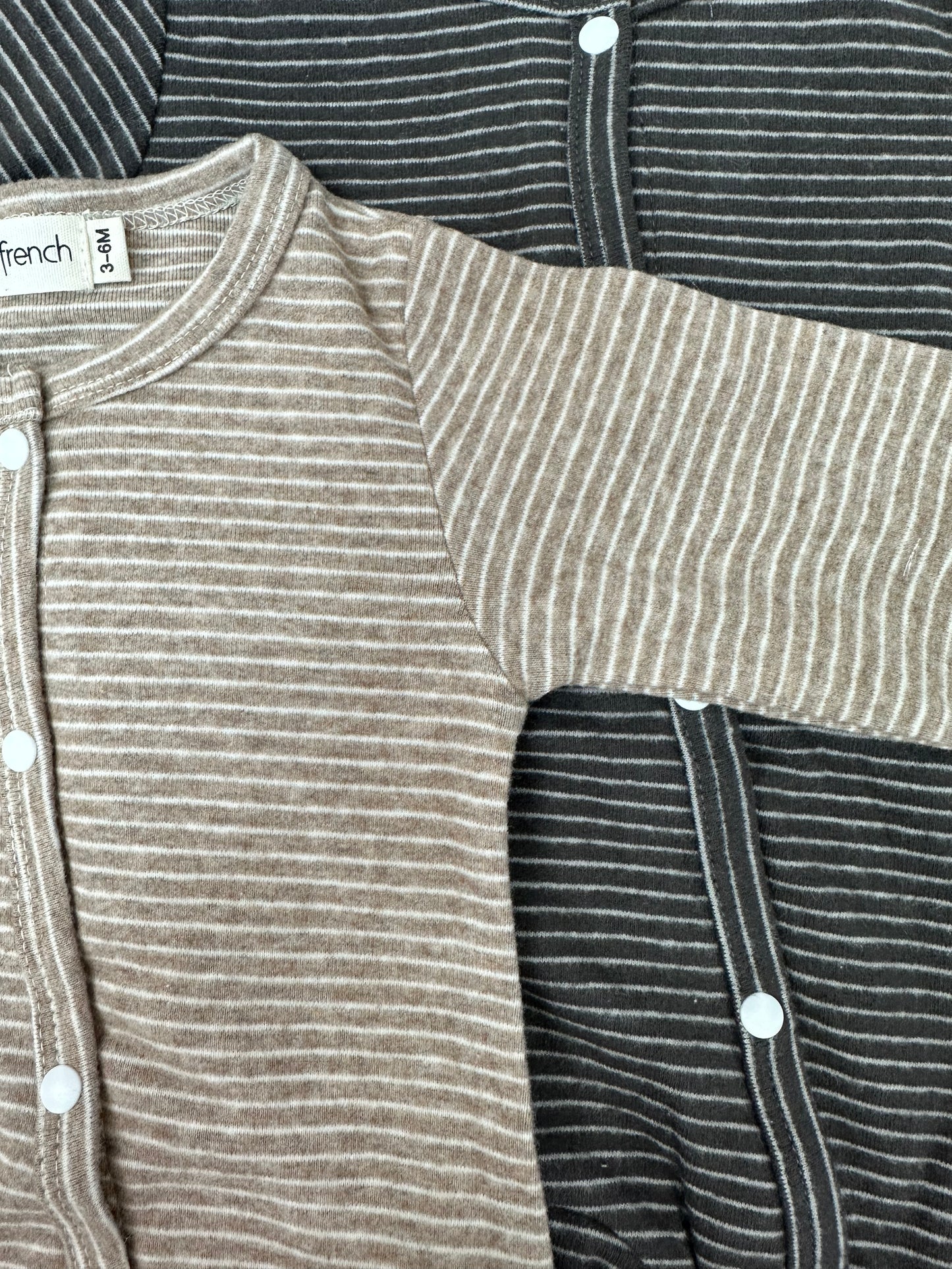 Two forever french baby Striped Snap Footie Pajamas are laid out overlapping—one beige with thin white stripes and one dark gray with matching white stripes, both featuring white snap closures.