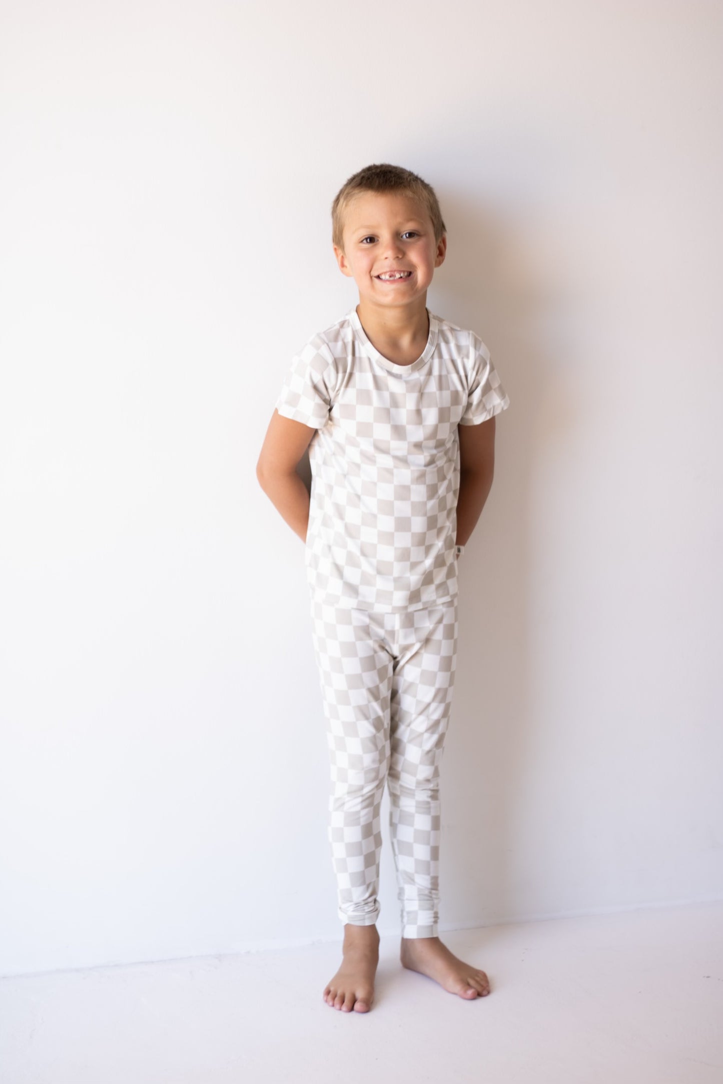 A young child smiles barefoot against a white wall, hands behind their back, wearing the forever french baby Bamboo Two Piece Lounge Set | Checkerboard in light colors.