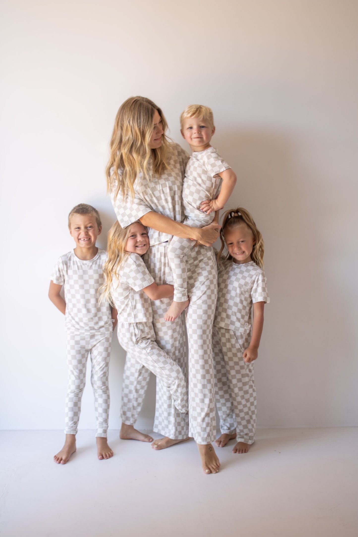 A woman and four young children smile together in matching forever french baby Bamboo Two Piece Lounge Set | Checkerboard pajamas, standing before a plain off-white wall.