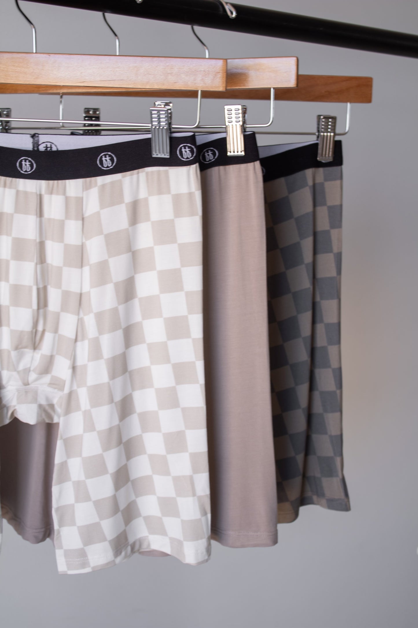 Three pairs of forever french baby Men's Bamboo Boxers (3 Pack) in Checkerboard & Faded Brown Checkerboard hang on clips from a rack against a plain background—hypo-allergenic essentials that combine comfort and style.
