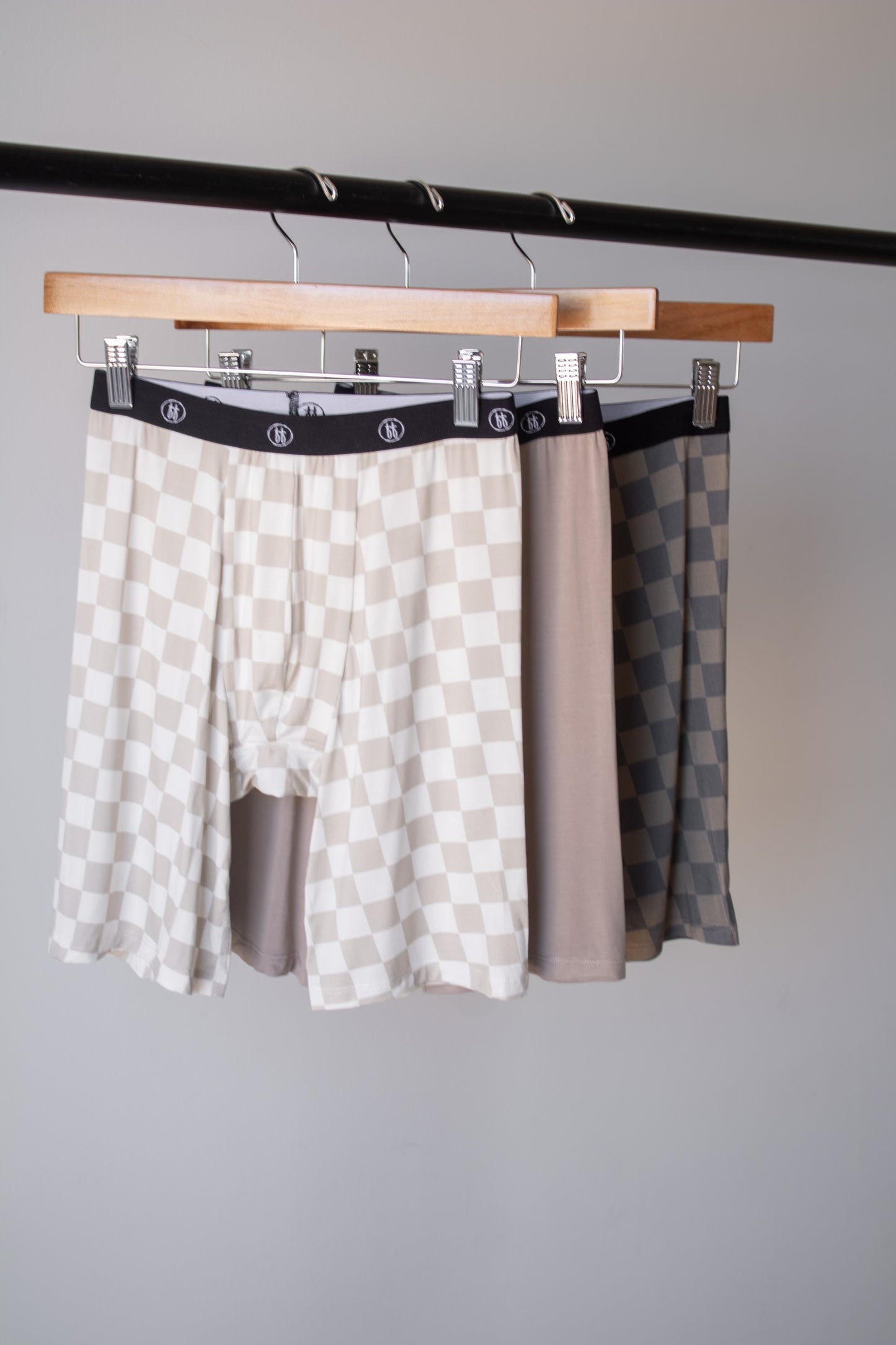 Three pairs of forever french baby Men's Bamboo Boxers (3 Pack) in checkerboard and faded brown checkerboard patterns hang on wooden hangers, shown against a plain light gray background. Hypo-allergenic and stylish in neutral tones.