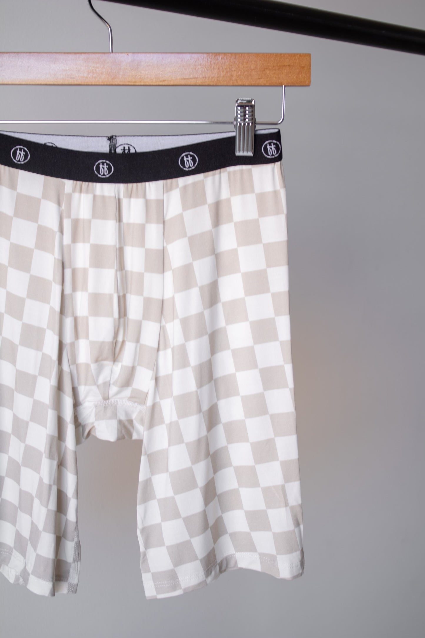 A pair of beige and white checkerboard Men’s Bamboo Boxers from forever french baby hangs on a wooden hanger against a light gray background. Features a black logo waistband—stylish, hypo-allergenic, and sold in a 3 pack.