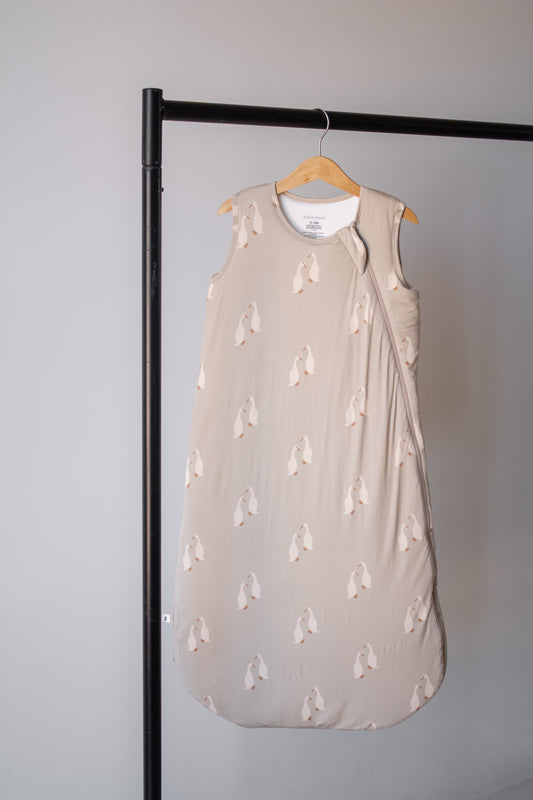 The Bamboo Sleep Sack | Silly Goose by forever french baby, featuring a beige penguin pattern, hangs on a wooden hanger from a black metal clothing rack against a plain light grey wall.