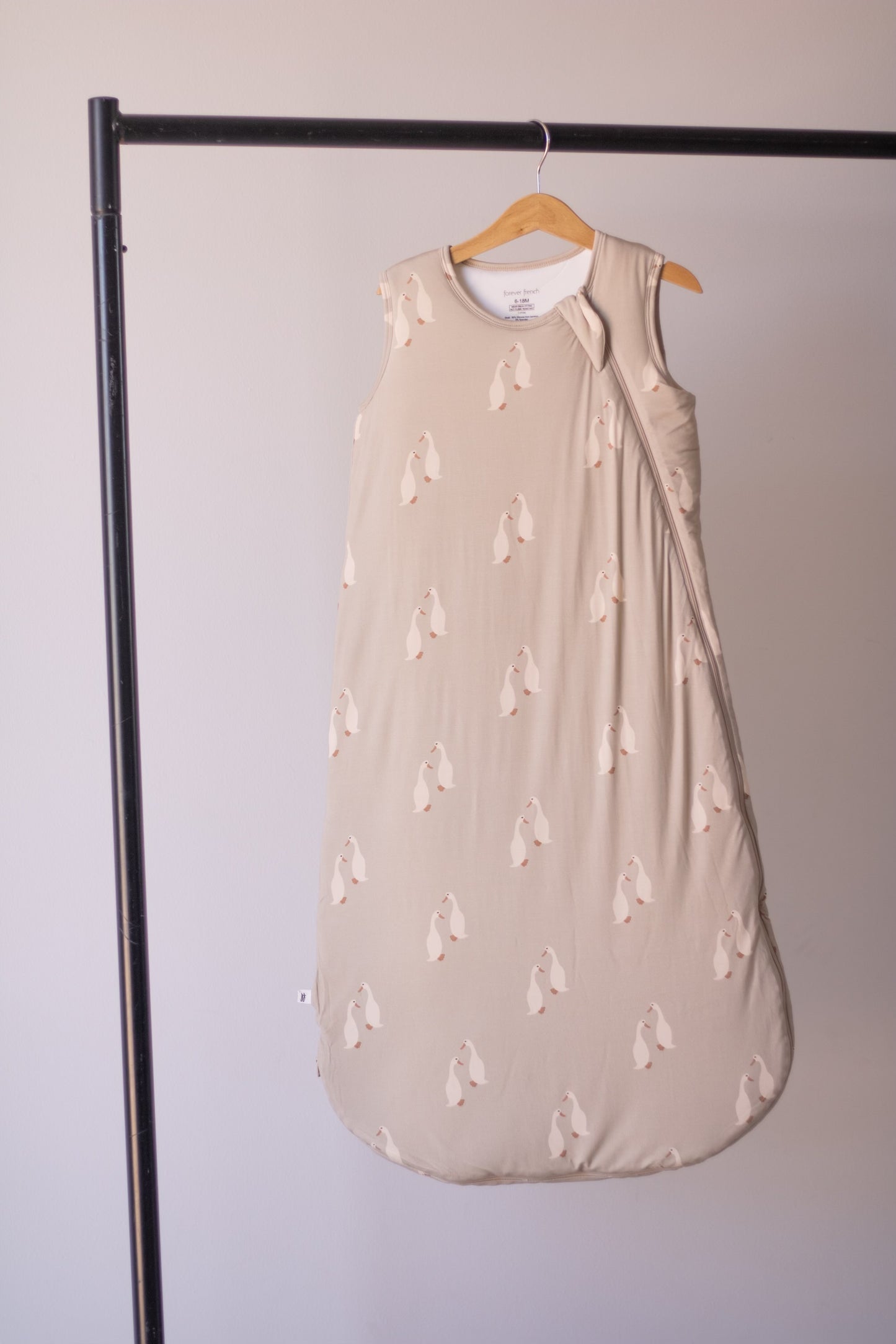 A beige Bamboo Sleep Sack | Silly Goose by forever french baby, featuring a penguin pattern, hangs sleeveless on a wooden hanger from a black metal rack against a plain light wall.