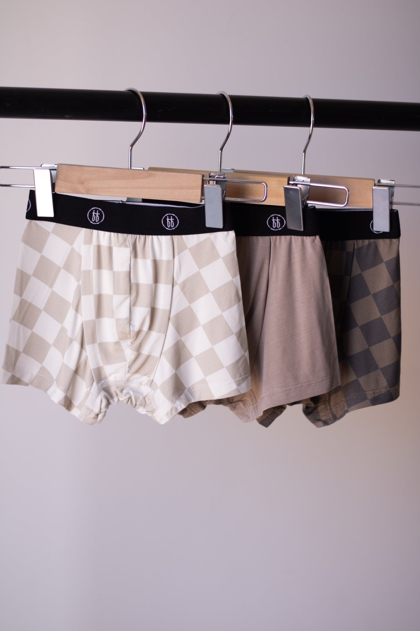 Three pairs of Bamboo Boxers (3 Pack) by forever french baby—Checkerboard and Faded Brown Checkerboard—hang from wooden clip hangers on a black rack. Enjoy hypo-allergenic comfort in stylish, neutral tones.